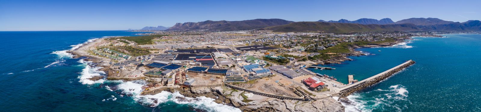Luftaufnahme des New Harbour von Hermanus mit Hafenmauer, Fischereigebäuden und der Stadt vor den Bergen